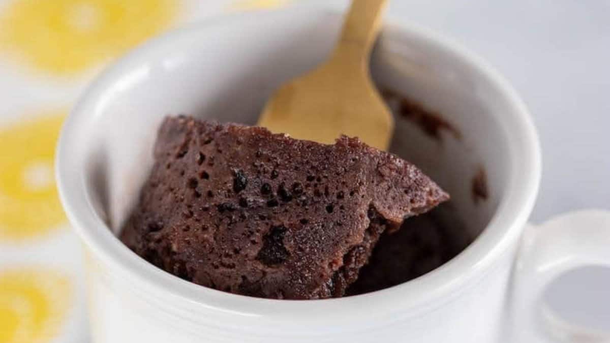 A close-up of a chocolate mug cake in a white mug, with a wooden spoon scooping out a portion. The cake looks moist and fluffy, sitting on a white surface with a yellow patterned background.