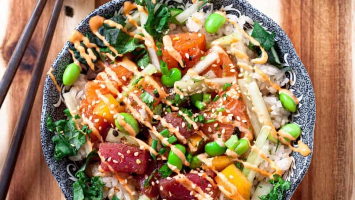 A colorful poke bowl topped with cubed raw fish, edamame, chopped greens, cucumber, mango, sesame seeds, and drizzled with orange sauce, served over rice in a patterned bowl with chopsticks on the side.