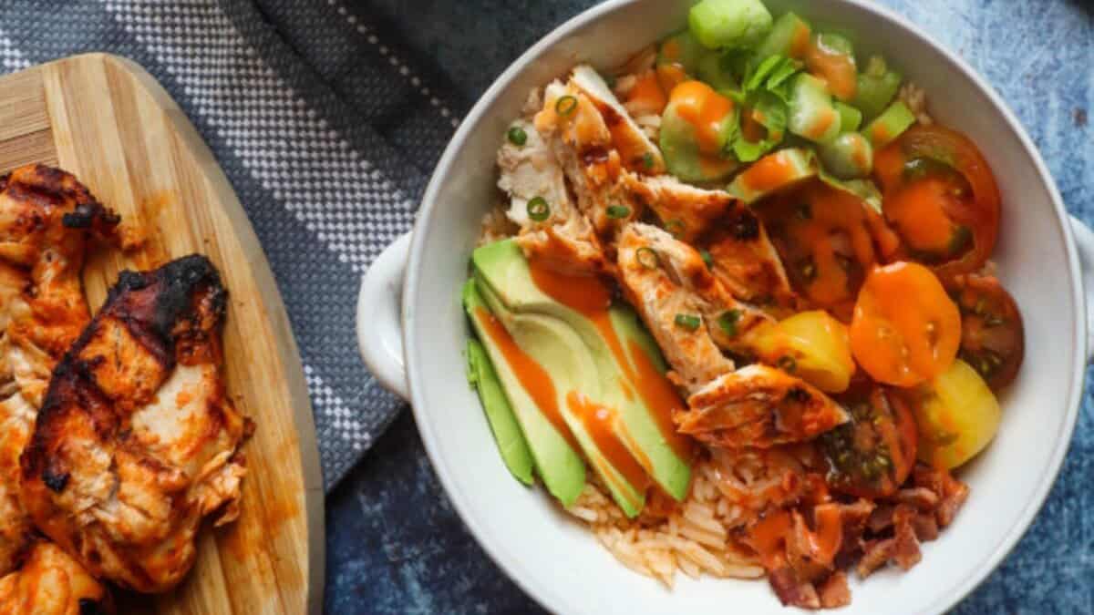 A bowl filled with sliced grilled chicken, avocado, cherry tomatoes, green onions, and sauce over rice. Beside the bowl, grilled chicken pieces rest on a wooden board. A blue-grey textured surface and cloth are in the background.