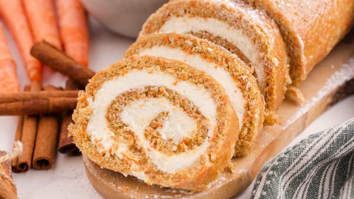 A sliced carrot cake roll with cream cheese filling is displayed on a wooden board, with fresh carrots and cinnamon sticks in the background.