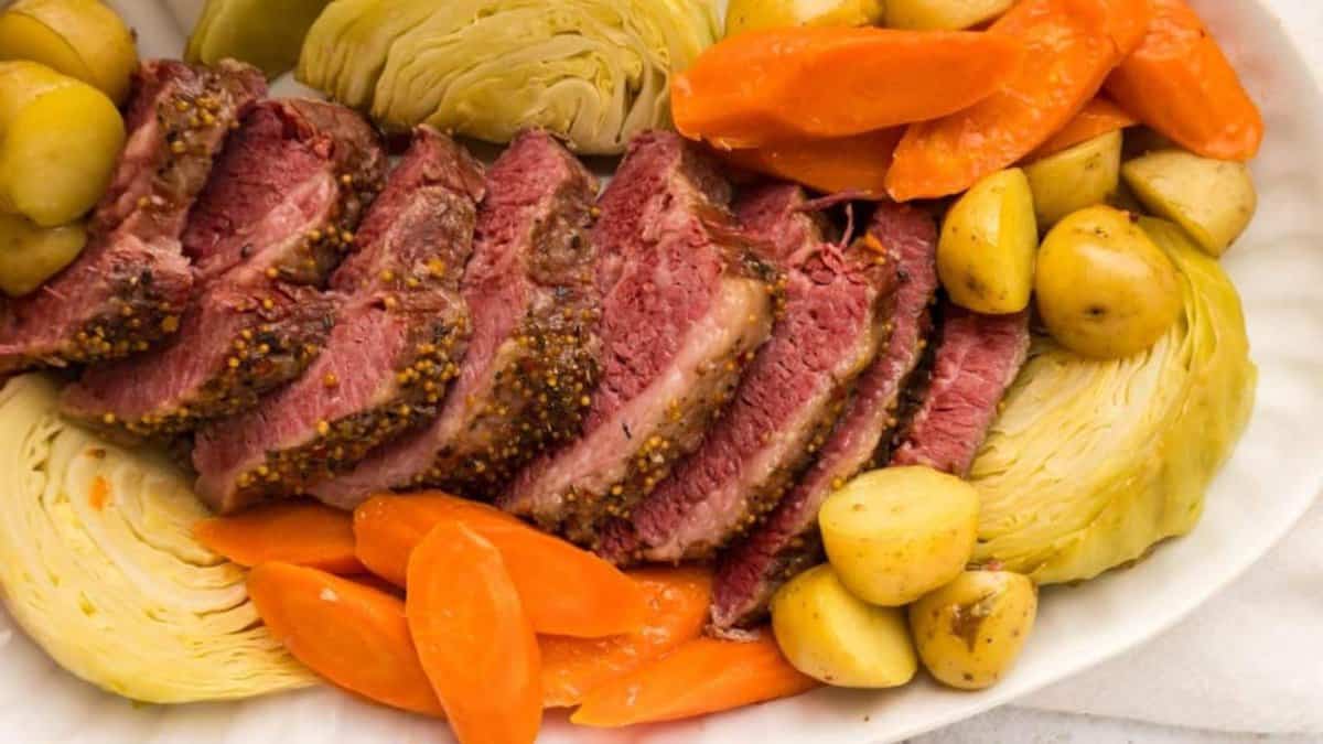 Sliced corned beef with a mustard glaze, surrounded by wedges of cabbage, sliced carrots, and small yellow potatoes on a white platter.
