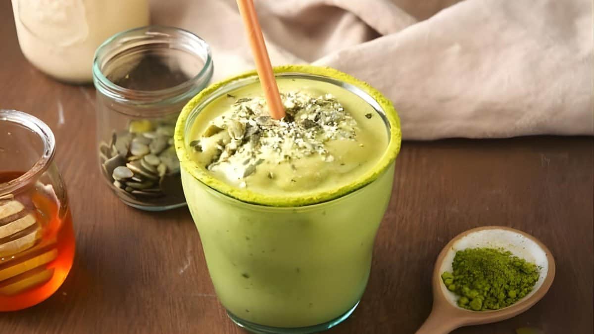 A glass of green matcha smoothie with a straw, topped with seeds. Nearby are a jar of pumpkin seeds, honey in a glass container, a spoonful of matcha powder, and a beige cloth on a wooden surface.