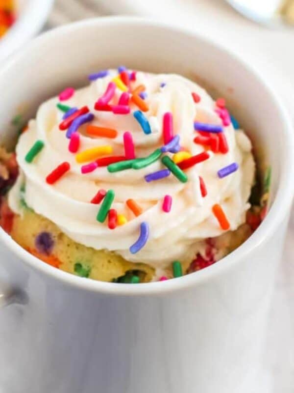 A white mug filled with a colorful confetti mug cake, topped with whipped cream and rainbow sprinkles, sits on a light wooden surface next to a small bowl of sprinkles and a spoon.