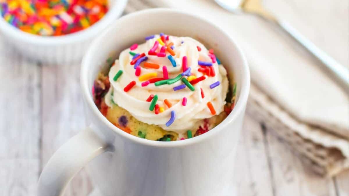A white mug filled with a colorful confetti mug cake, topped with whipped cream and rainbow sprinkles, sits on a light wooden surface next to a small bowl of sprinkles and a spoon.