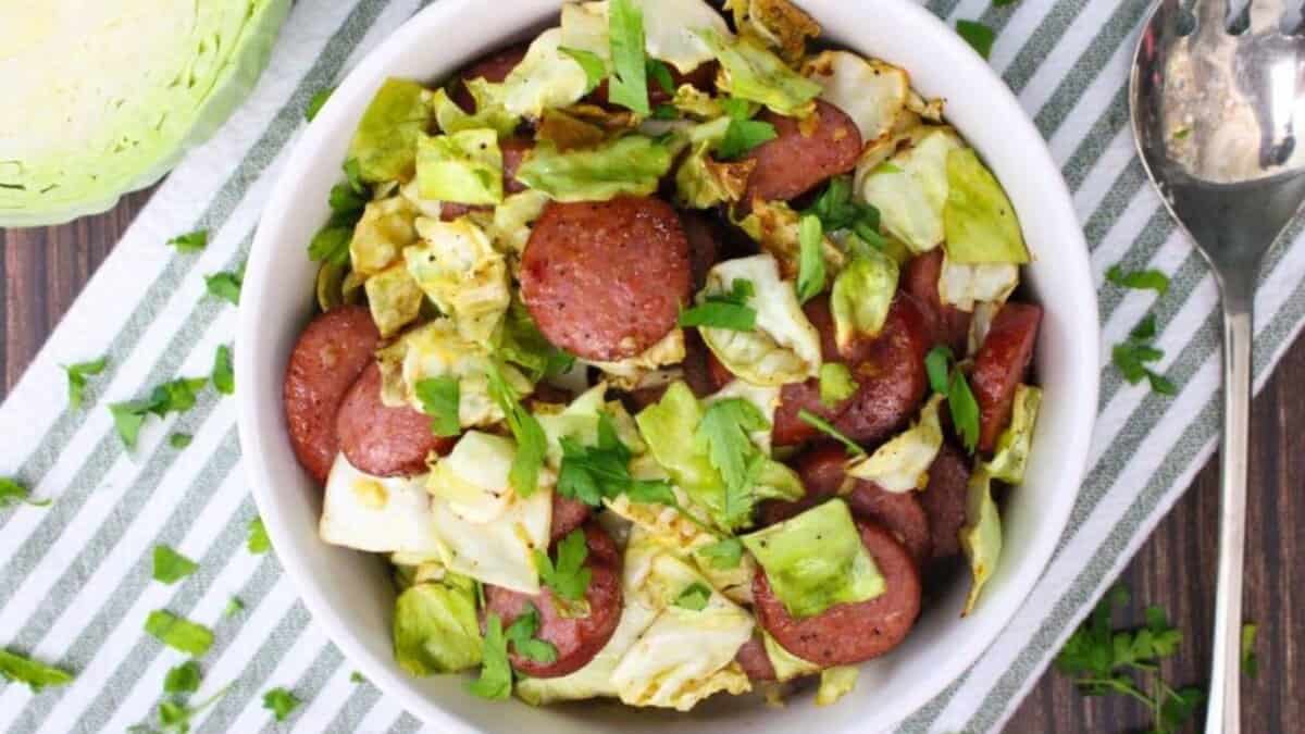 A white bowl filled with sliced sausage, cooked cabbage, and sprinkled fresh parsley, placed on a striped cloth with a spoon and cabbage on the side.