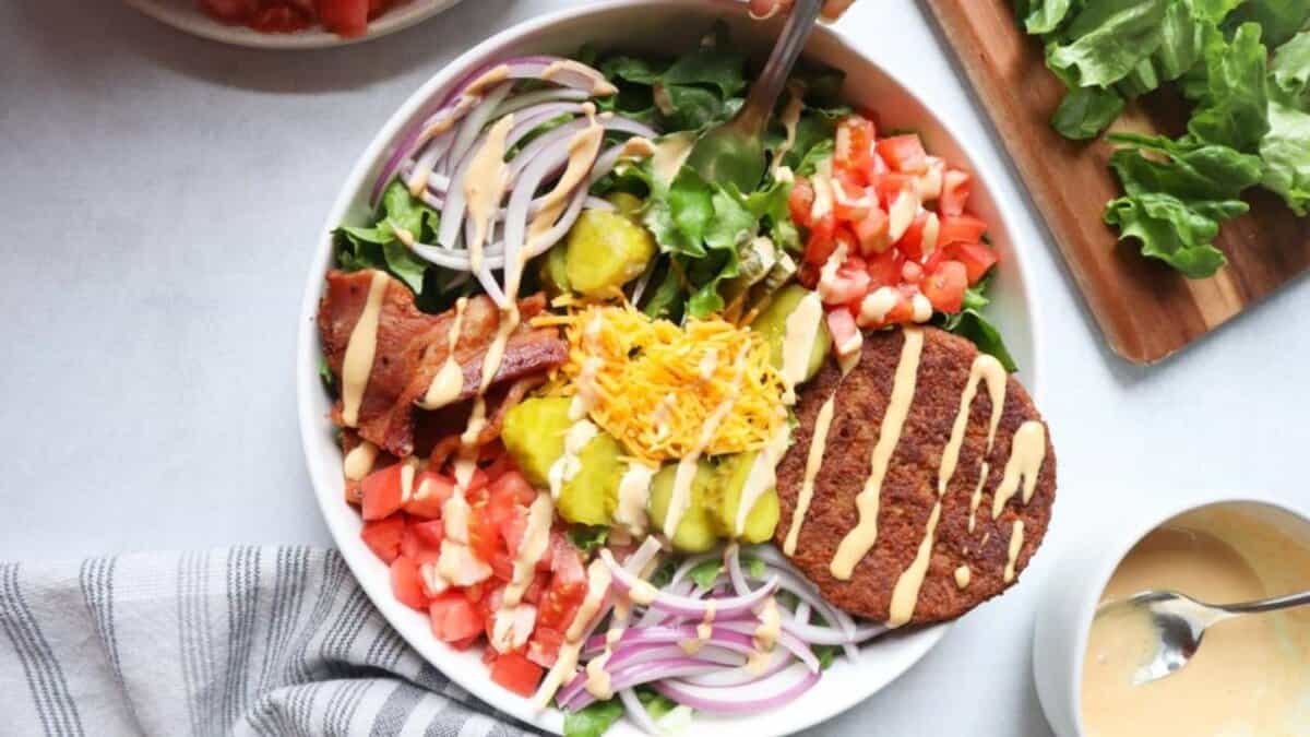 A bowl filled with leafy greens, sliced tomatoes, red onions, pickles, bacon, shredded cheese, and a burger patty, topped with creamy dressing. A fork is in the bowl, and other ingredients are nearby.