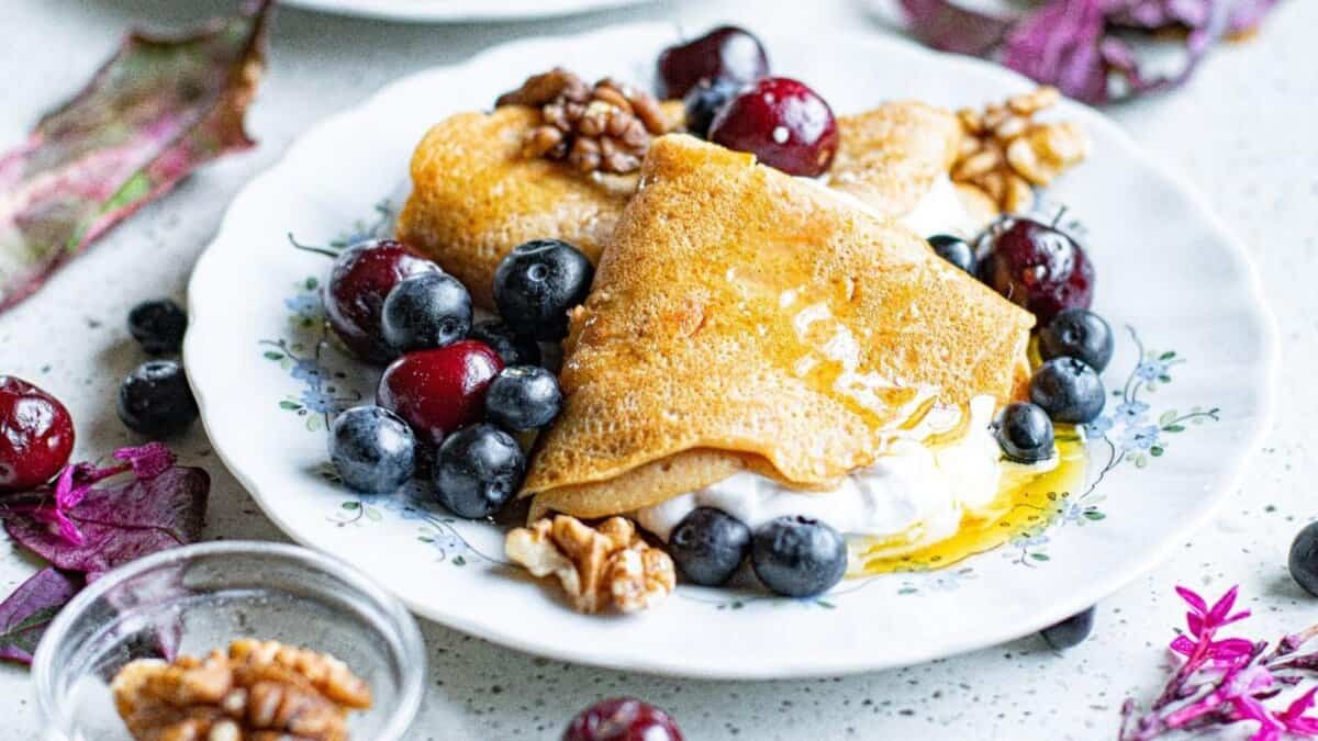 A plate with folded crepes topped with honey, walnuts, blueberries, grapes, and a dollop of cream, garnished with leaves and surrounded by scattered berries and nuts.