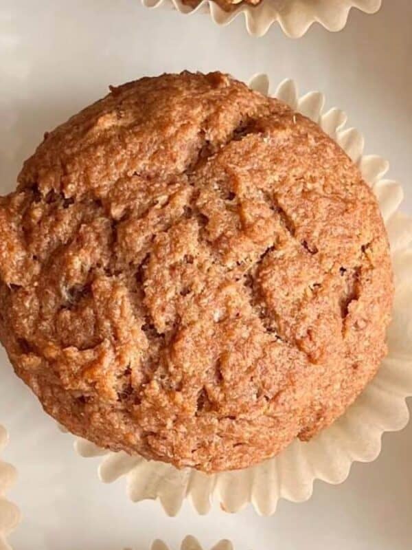 A close-up view of several brown muffins in white paper liners arranged on a white surface. The muffins have a textured, slightly cracked top.