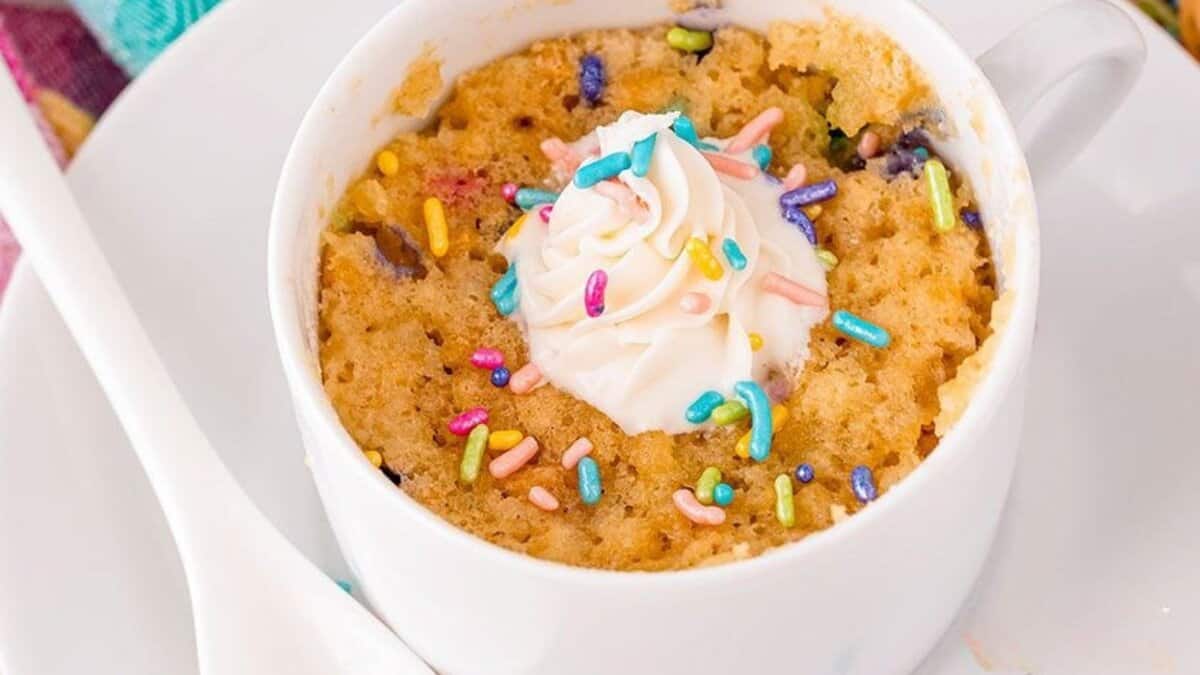 A white mug filled with a confetti mug cake, topped with a swirl of whipped cream and colorful sprinkles. A white spoon rests beside the mug on a white plate.