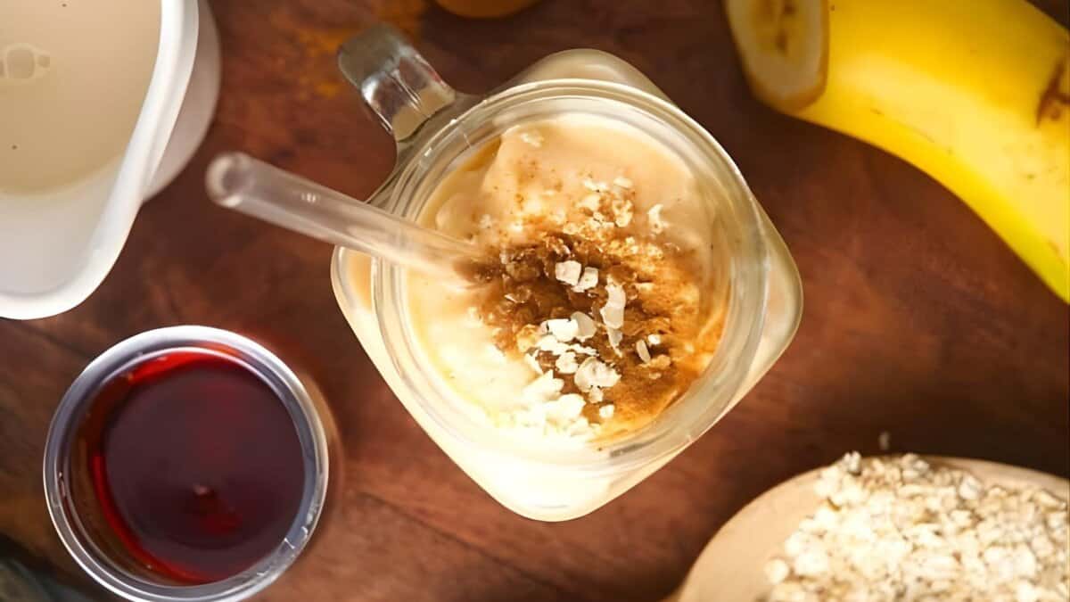 A glass mug with a smoothie topped with oats and cinnamon, a clear straw, surrounded by a banana, a cup of red liquid, and a wooden spoon with more oats on a wooden surface.
