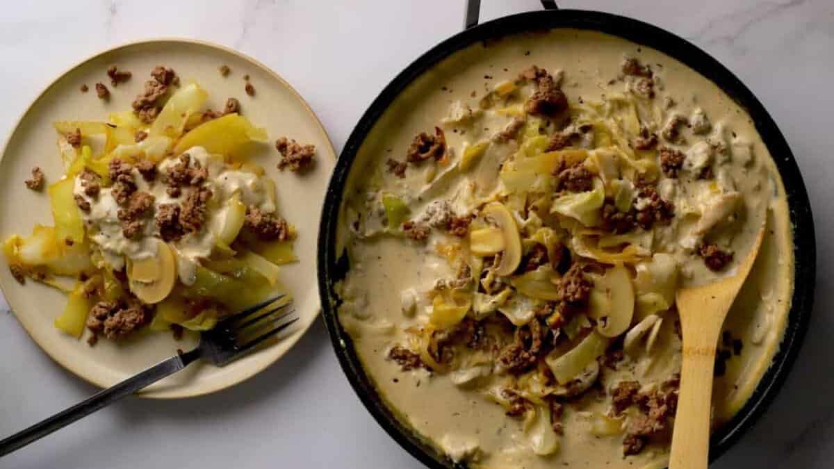 A skillet filled with creamy ground beef, mushrooms, onions, and cabbage sits beside a plate with a serving of the same dish. A fork and knife rest on the plate.