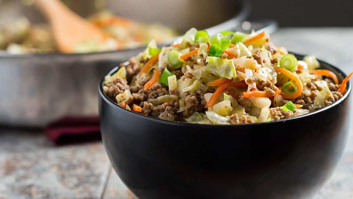 A black bowl filled with a mixture of ground meat, chopped cabbage, carrots, and sliced green onions sits on a table, with a pot and wooden spoon blurred in the background.