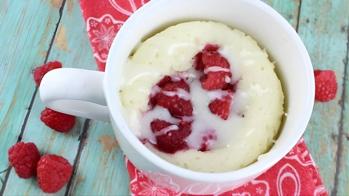 A white mug filled with a baked raspberry mug cake topped with icing, placed on a red patterned napkin with fresh raspberries scattered on a rustic blue wooden surface.