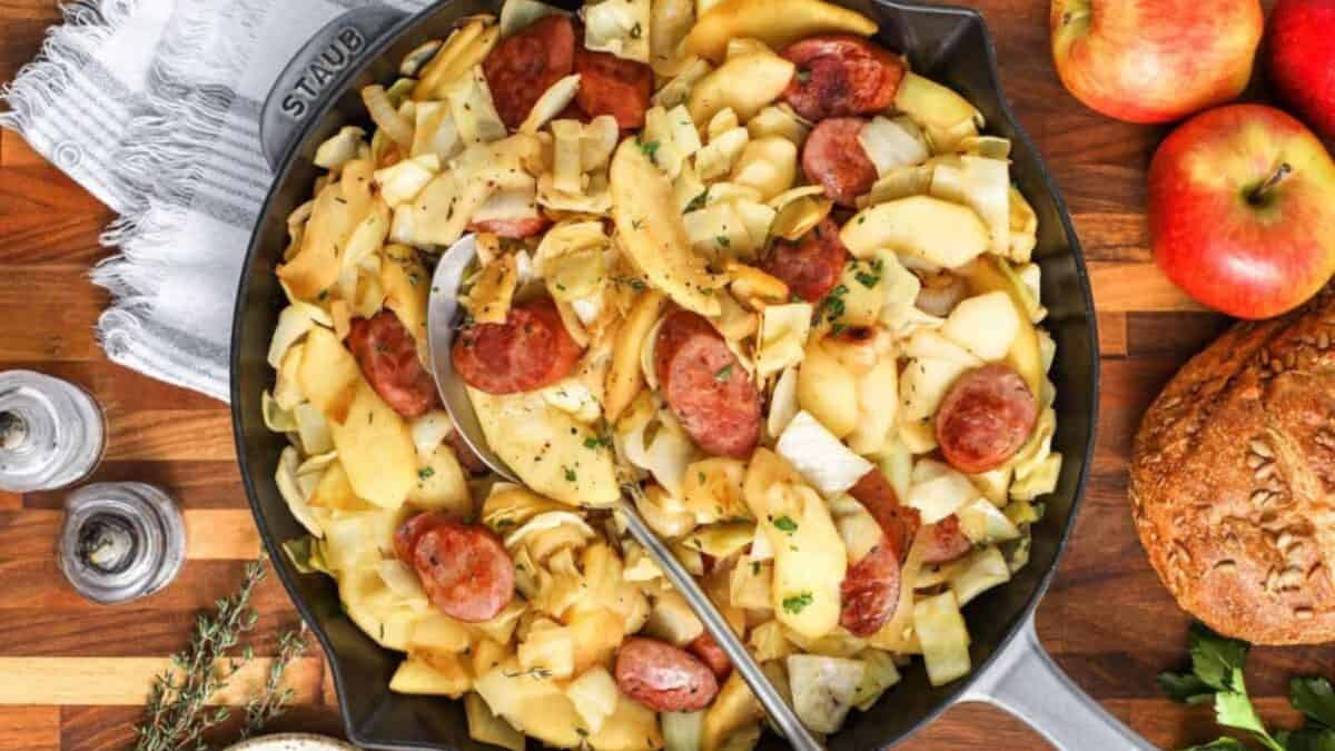 A skillet filled with sliced sausage, cabbage, and apples, garnished with herbs. A spoon rests in the skillet. Nearby are whole apples, a loaf of bread, salt and pepper shakers, and a sprig of thyme on a wooden surface.