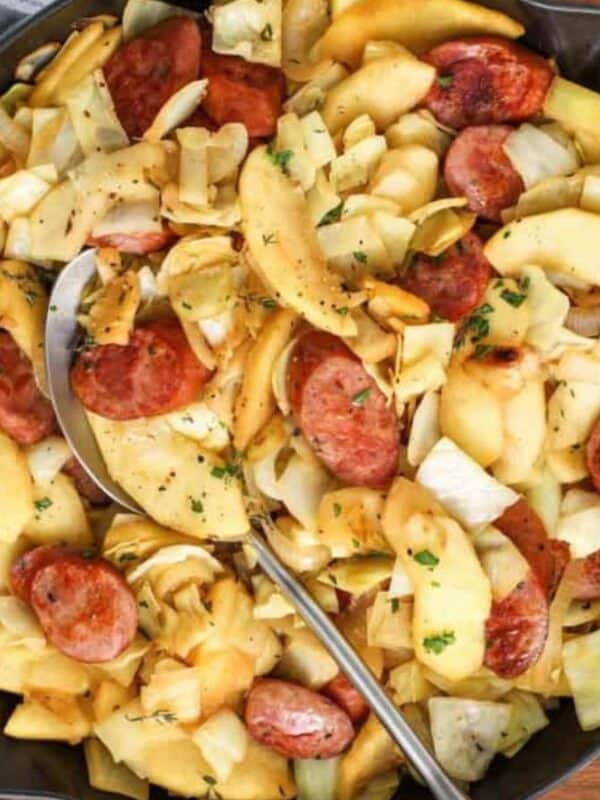 A skillet filled with sliced sausage, cabbage, and apples, garnished with herbs. A spoon rests in the skillet. Nearby are whole apples, a loaf of bread, salt and pepper shakers, and a sprig of thyme on a wooden surface.