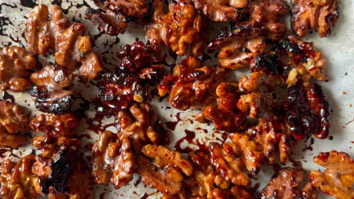 Close-up of caramelized walnuts scattered on a baking sheet, coated in a shiny, dark glaze with some areas slightly burnt.