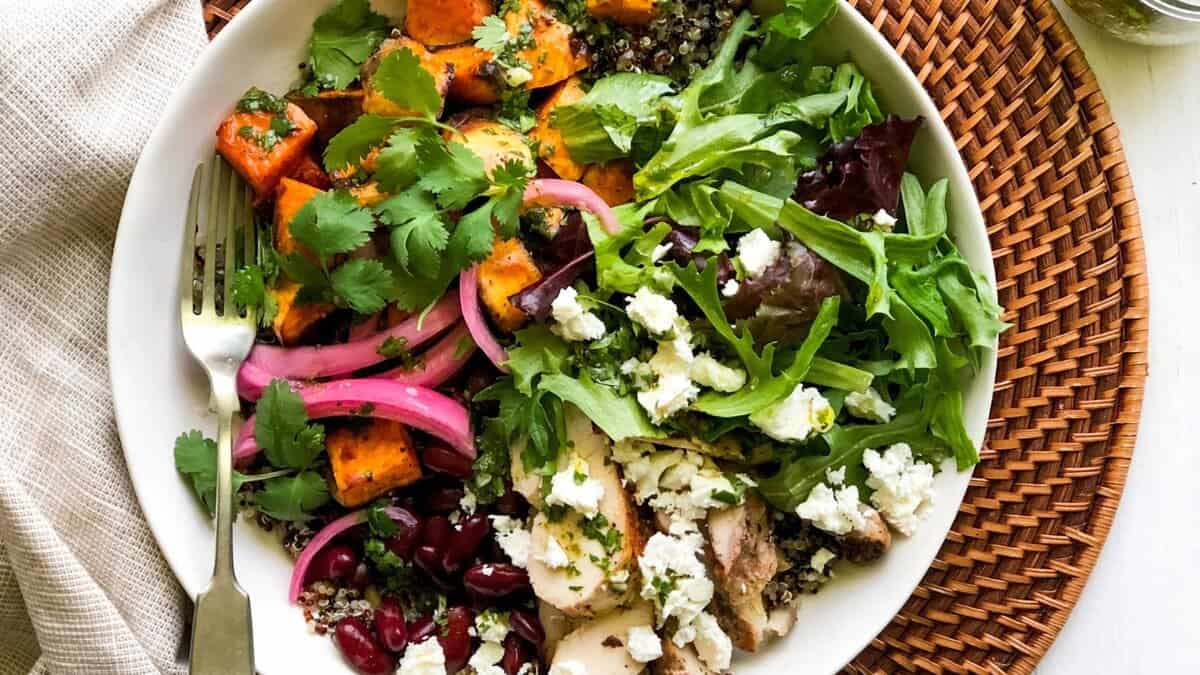 A bowl filled with mixed greens, crumbled feta, grilled chicken, kidney beans, roasted sweet potatoes, quinoa, pickled onions, and fresh cilantro, placed on a woven placemat with a fork on the side.