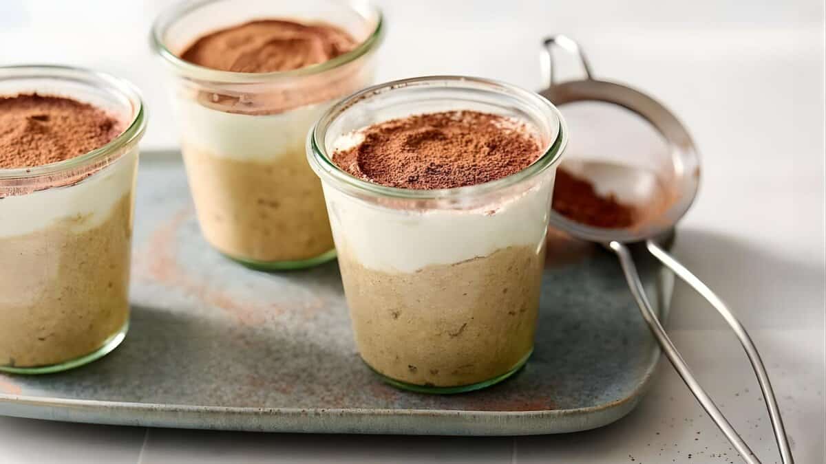 A hand holds a spoon in a glass jar filled with overnight oats, chia seeds, blueberries, and nuts. Another similar jar sits in the background, next to oats and blueberries in small bowls. A yellow-striped towel is nearby.