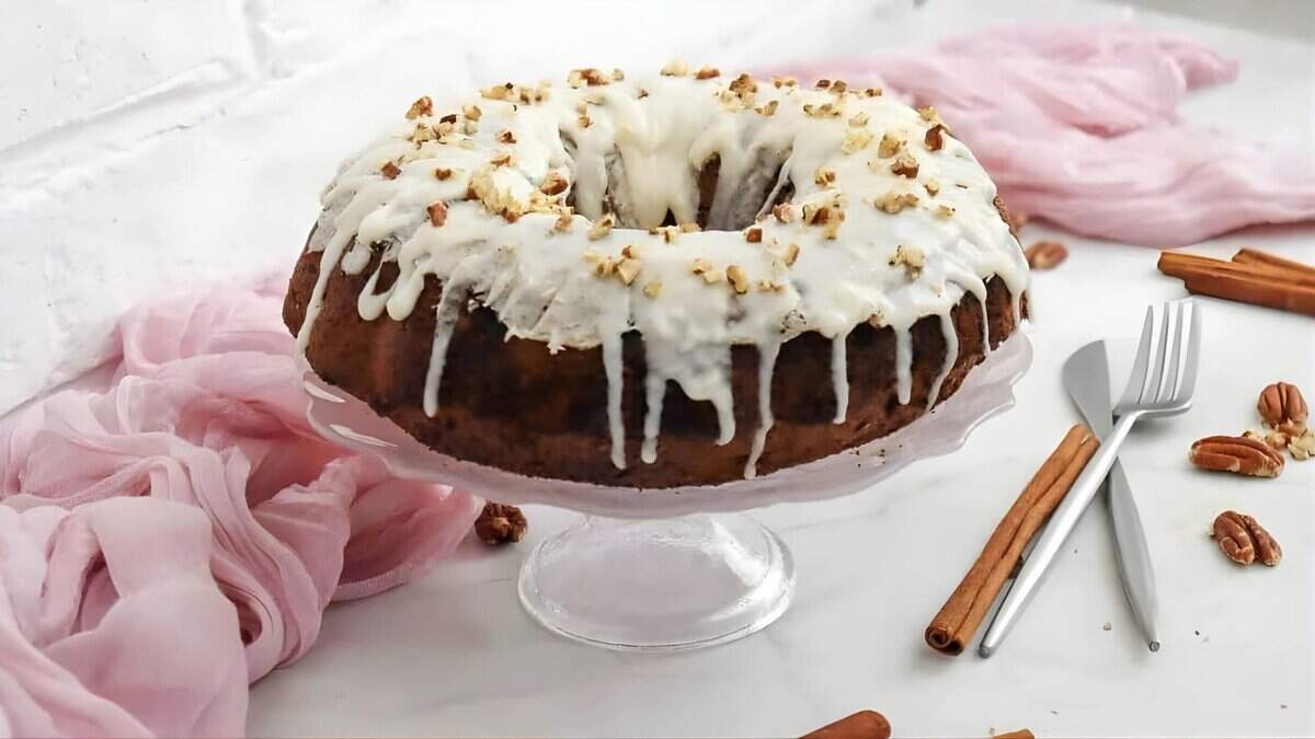 A bundt cake with white icing and chopped nuts on top sits on a glass stand. Forks, cinnamon sticks, pecans, and pink fabric decorate the white marble surface around it.