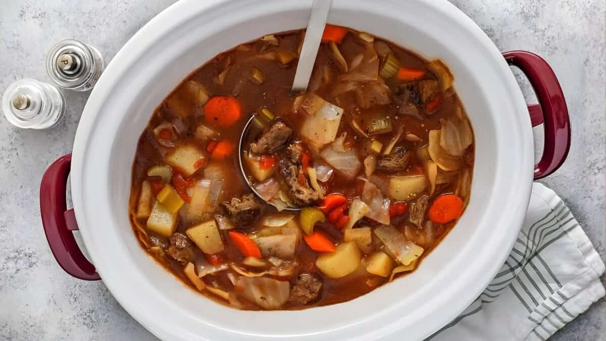 A white slow cooker filled with beef and vegetable stew, including chunks of beef, carrots, potatoes, celery, and cabbage in a brown broth. A ladle rests inside. A striped towel and salt and pepper shakers are nearby.