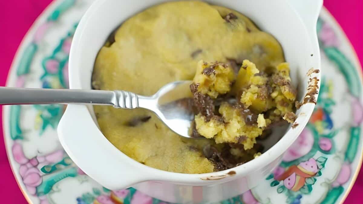 A spoon scooping out a portion of a chocolate chip cookie baked in a white ramekin, placed on a colorful floral-patterned plate with a pink background.