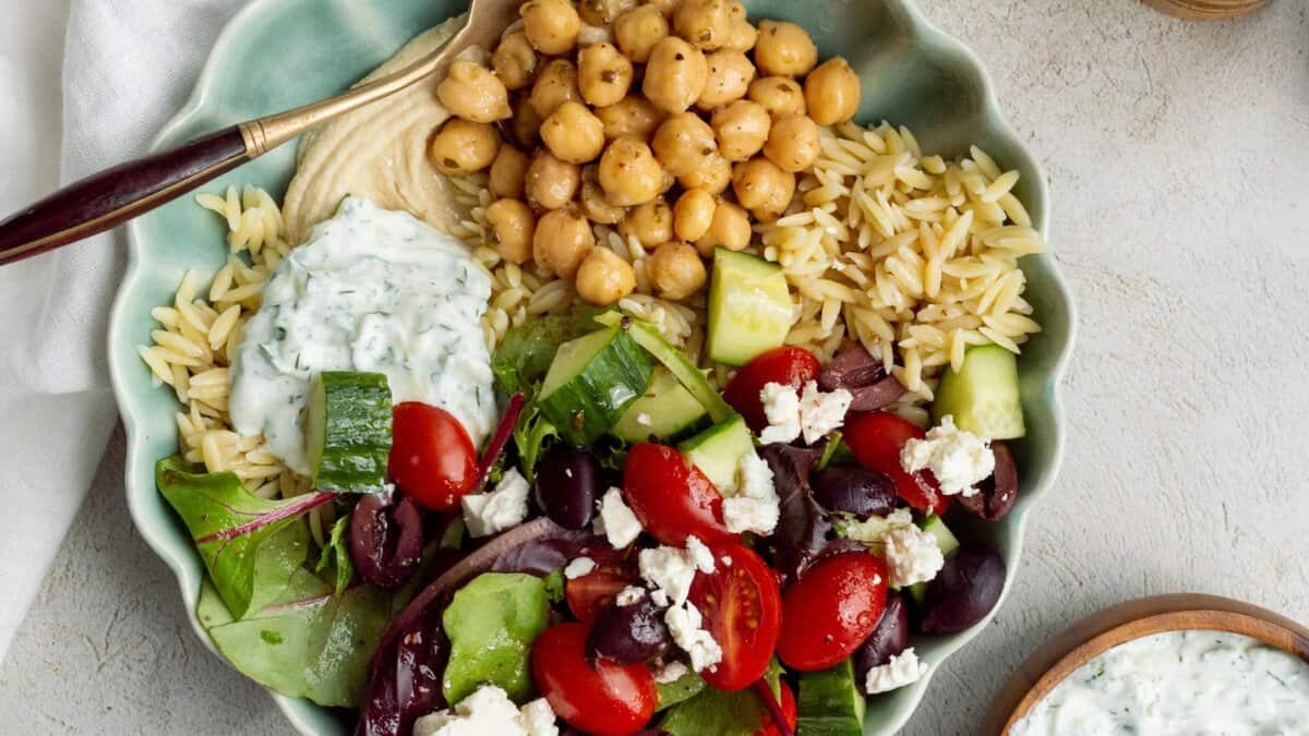 A bowl with orzo pasta, chickpeas, hummus, cucumber, tomatoes, feta cheese, olives, mixed greens, and tzatziki sauce, garnished with herbs and served with a fork.