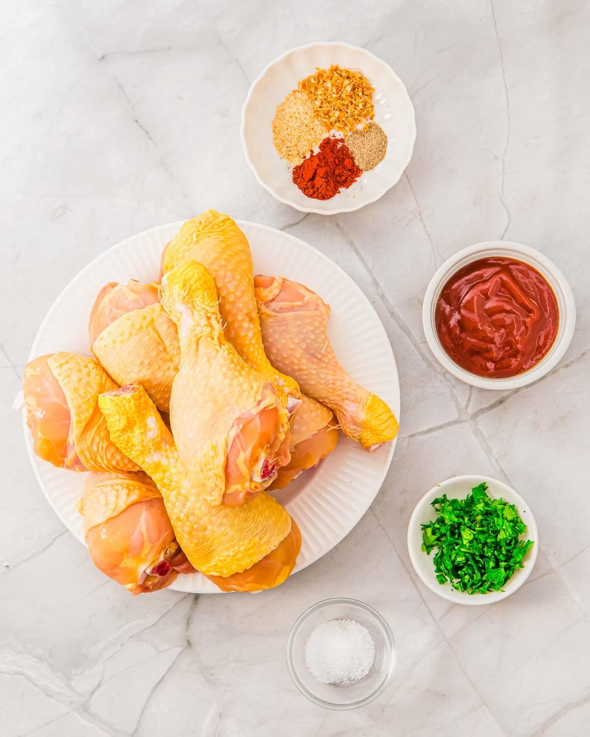 Raw chicken drumsticks on a white plate, surrounded by small bowls containing spices, BBQ sauce, chopped herbs, and salt, arranged on a white marble surface.
