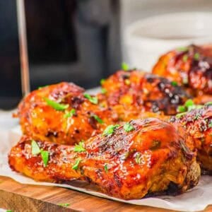 Close-up of BBQ glazed, air fryer chicken drumsticks garnished with chopped fresh herbs, arranged on a wooden board lined with parchment paper. The chicken has a shiny, caramelized surface.