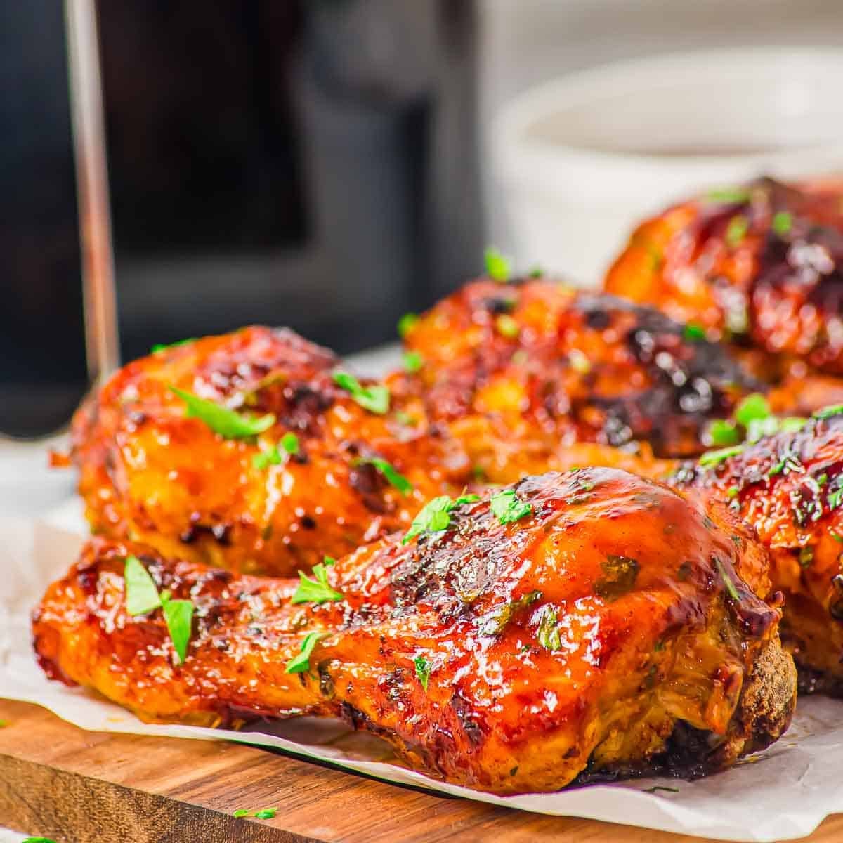 Close-up of BBQ glazed, air fryer chicken drumsticks garnished with chopped fresh herbs, arranged on a wooden board lined with parchment paper. The chicken has a shiny, caramelized surface.