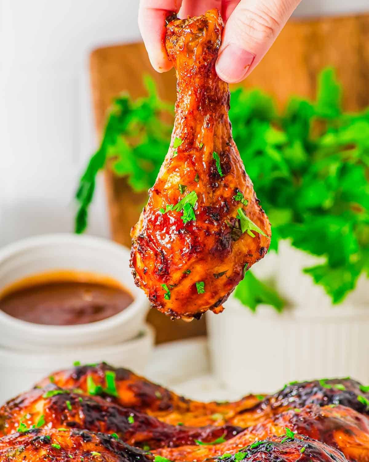 A hand holds up a glazed, crispy air fried chicken drumstick garnished with chopped herbs. In the background, there is a bowl of dipping sauce and a bunch of fresh parsley.