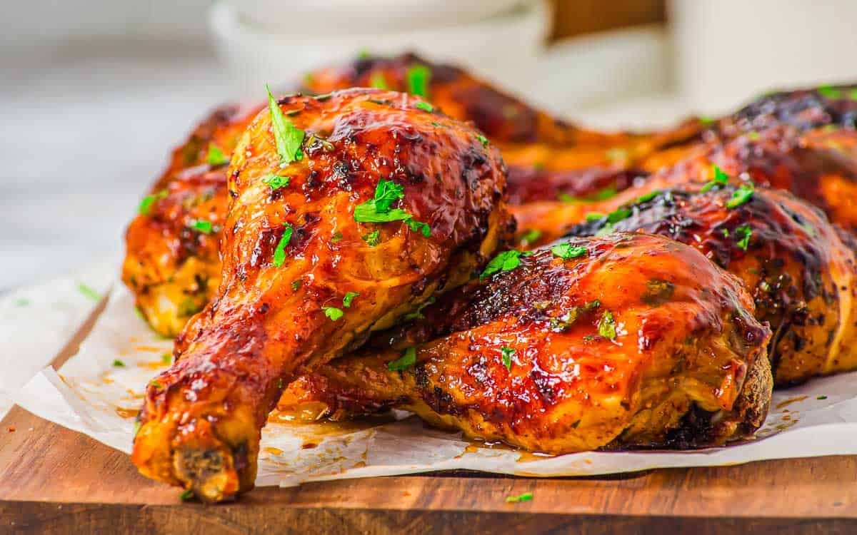 Barbecued air fryer chicken drumsticks and thighs with a glossy, caramelized glaze, garnished with chopped fresh herbs, served on parchment paper on a wooden board.