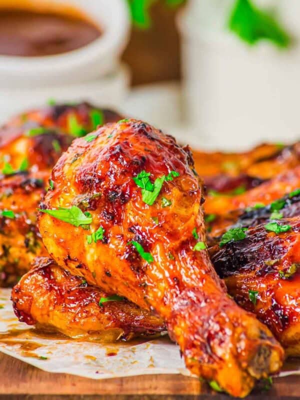 Close-up of glazed, chicken drumsticks garnished with chopped parsley, served on parchment paper with a cup of sauce in the background.