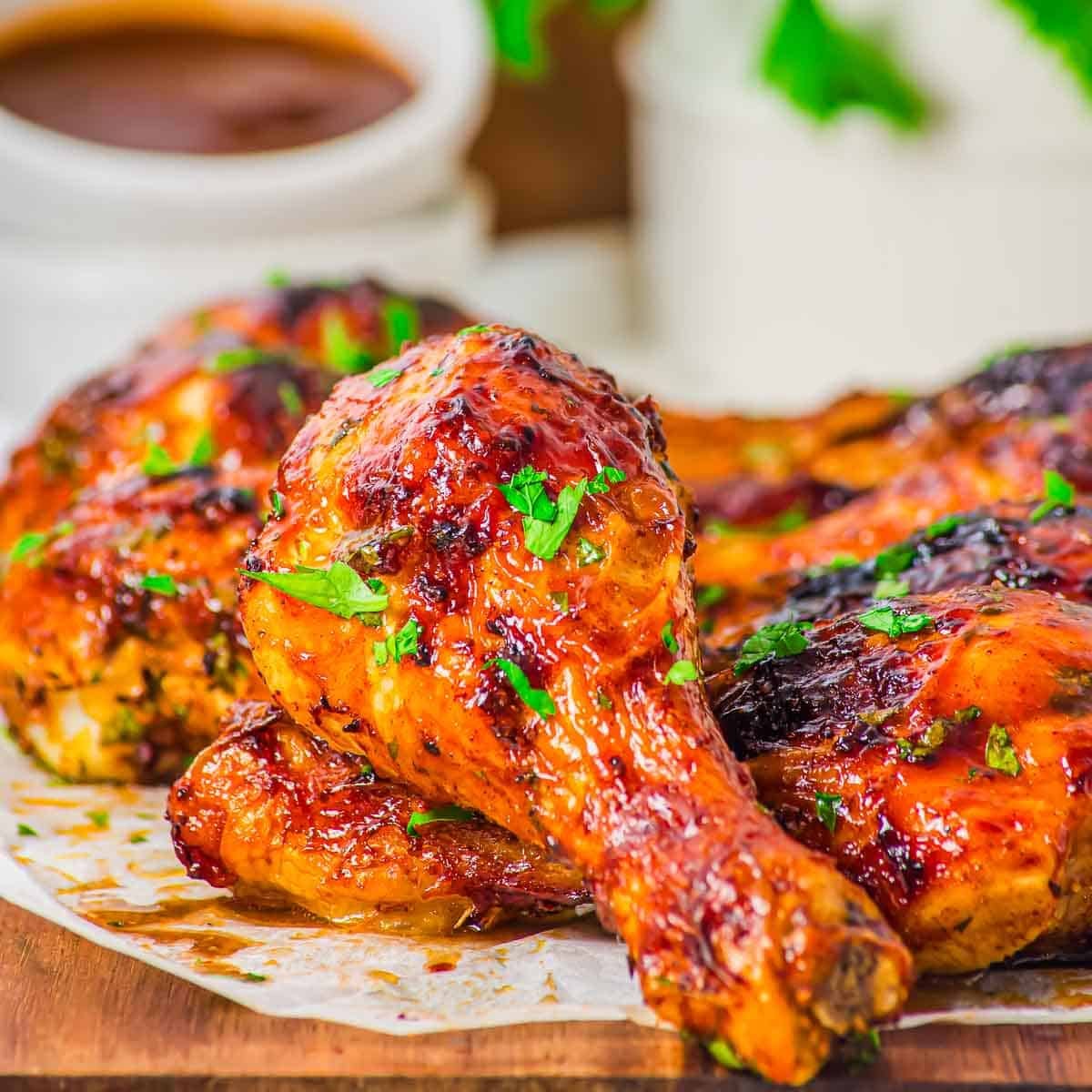 Close-up of glazed, chicken drumsticks garnished with chopped parsley, served on parchment paper with a cup of sauce in the background.