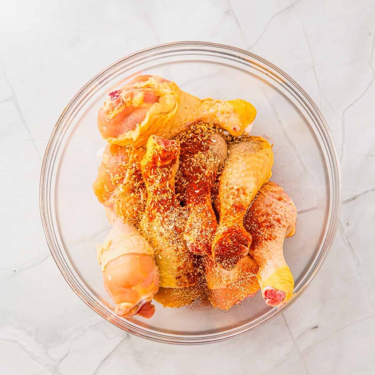 A glass bowl filled with raw chicken drumsticks sprinkled with spices, set on a white marble surface.