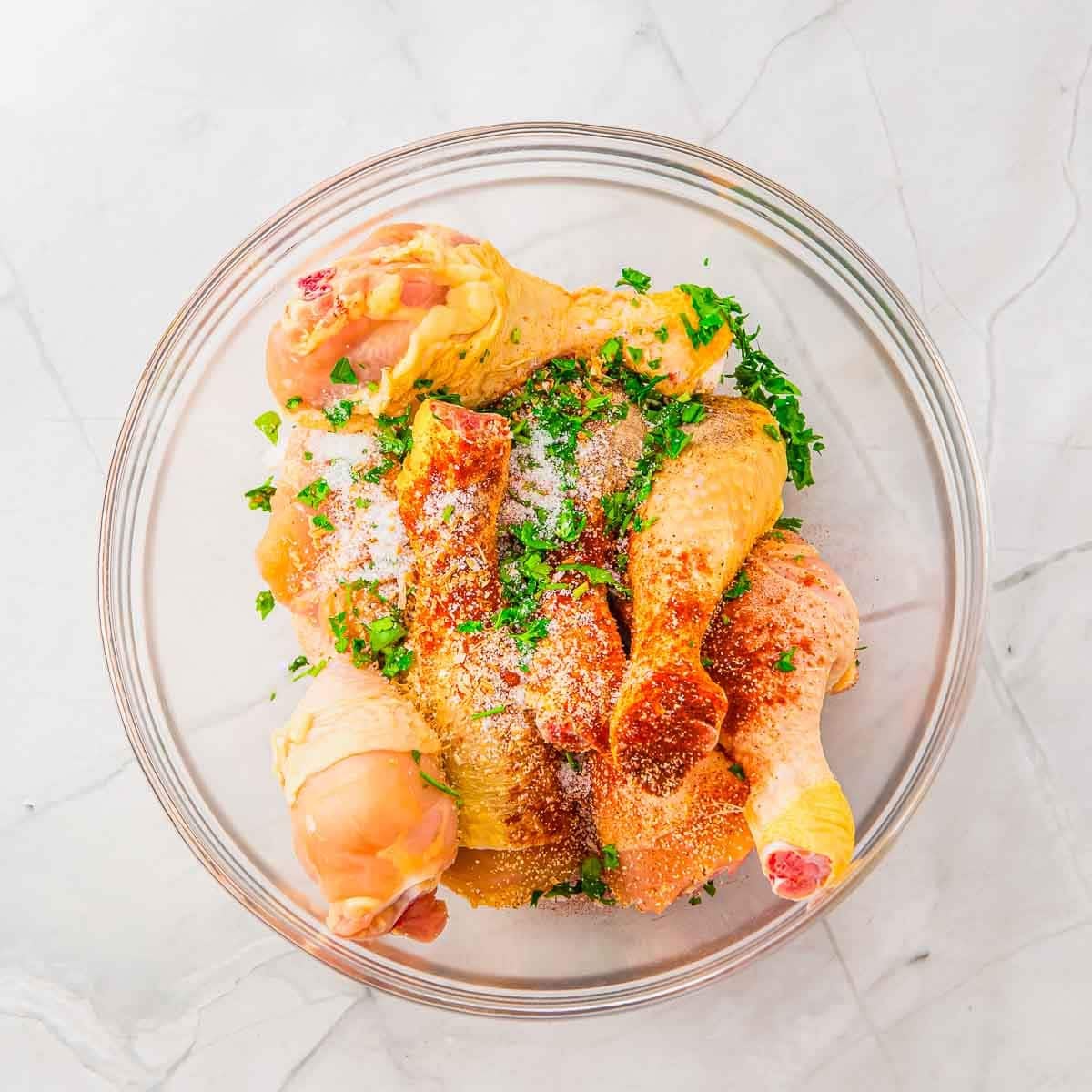 Raw chicken drumsticks in a glass bowl, sprinkled with chopped fresh herbs, salt, and seasoning, set on a light-colored marble surface.