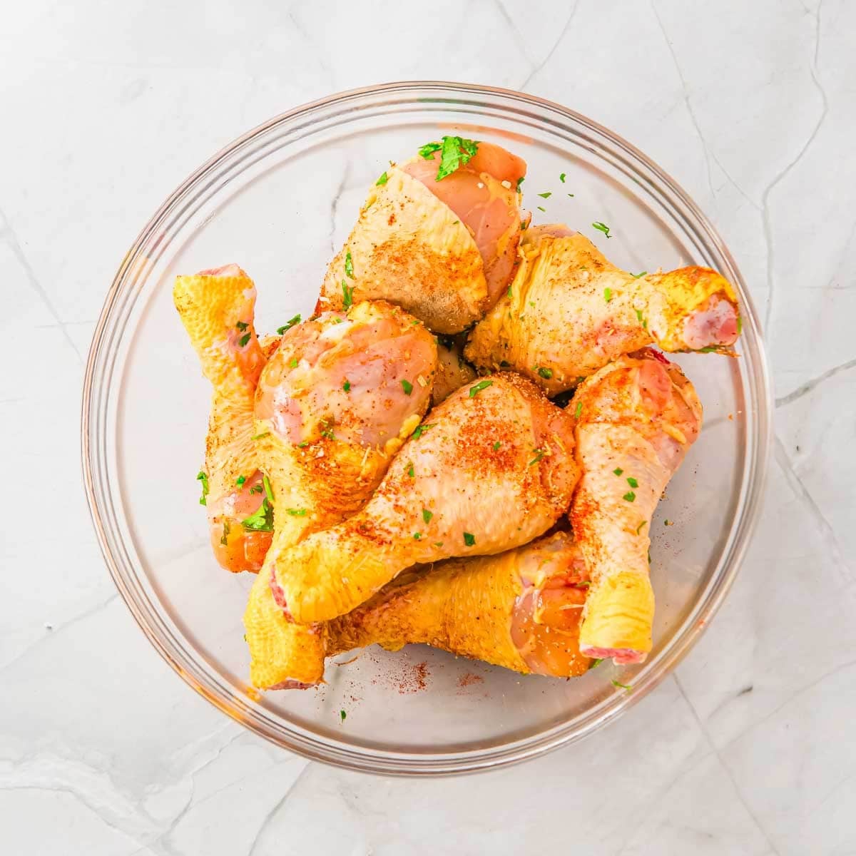 A glass bowl filled with raw chicken drumsticks, sprinkled with seasoning and chopped herbs, sits on a light-colored marble surface.