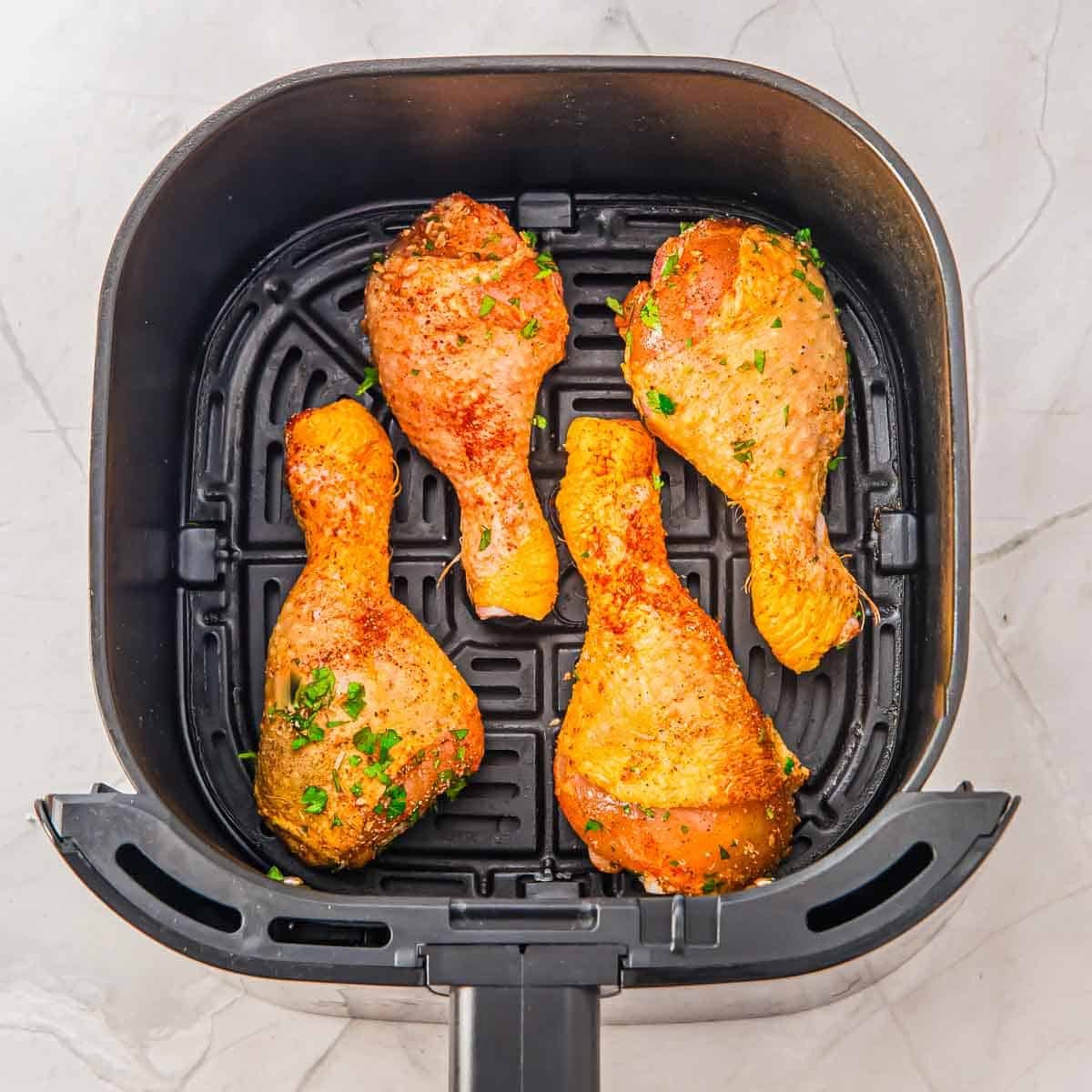 Four seasoned chicken drumsticks arranged in a single layer inside an air fryer basket on a light-colored countertop.