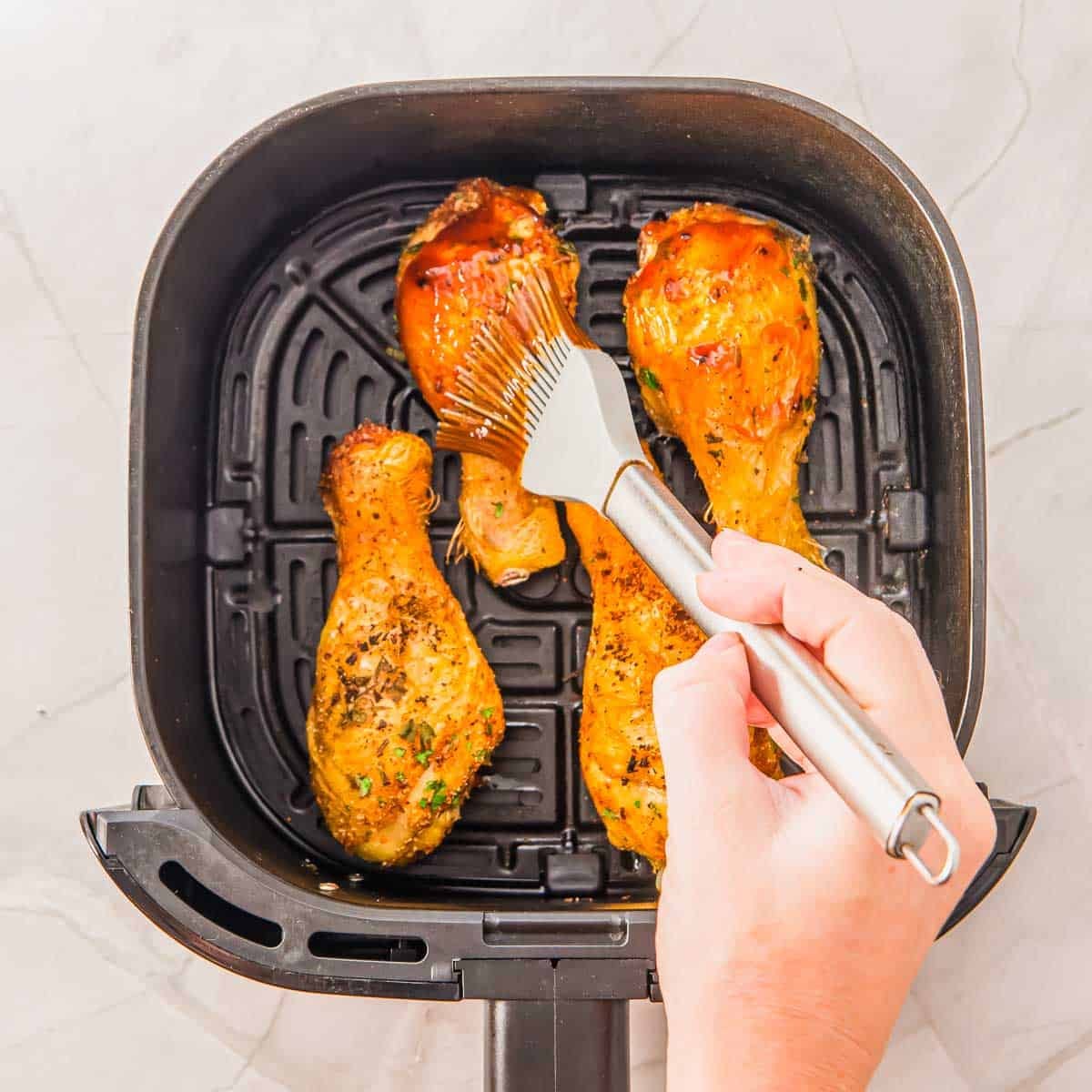 A hand uses a brush to apply BBQ sauce to cooked chicken drumsticks inside an air fryer basket on a light-colored surface.