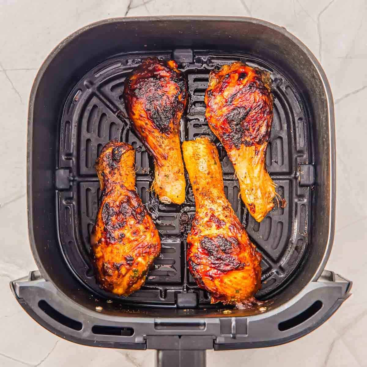 Four cooked, slightly charred chicken drumsticks in an air fryer basket on a light marble countertop.