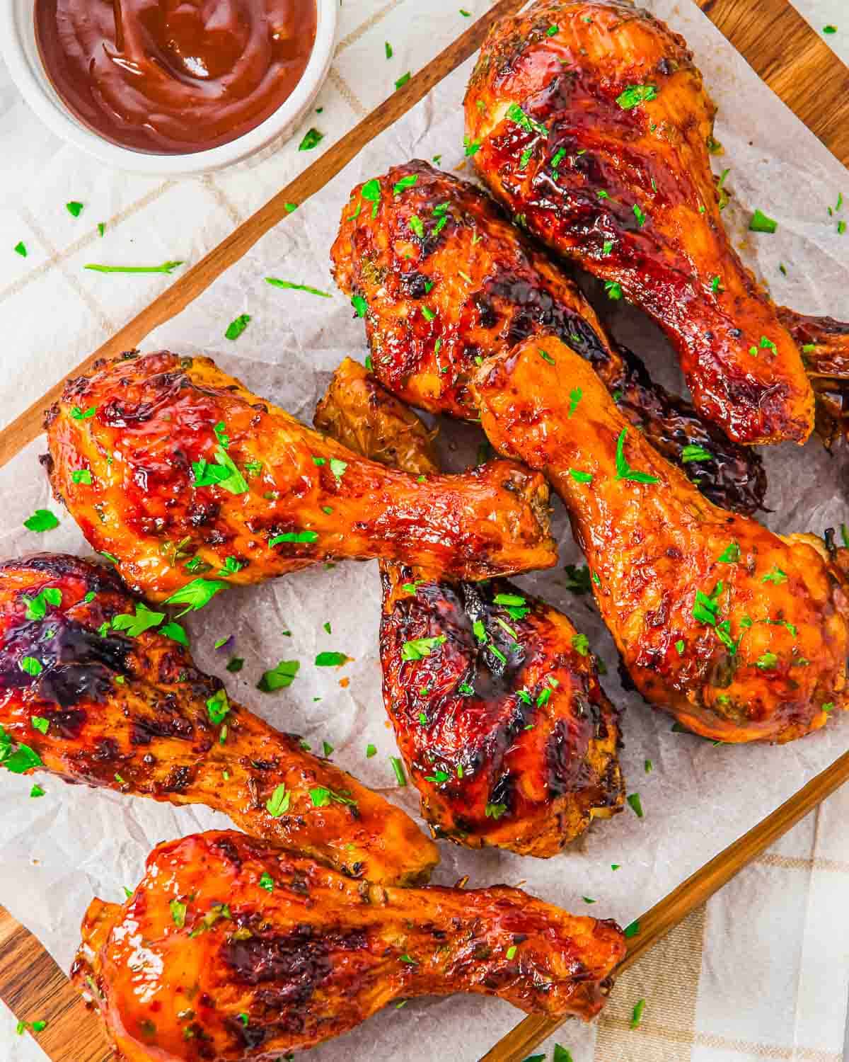 Air fried chicken drumsticks glazed with barbecue sauce, garnished with chopped parsley, arranged on parchment paper atop a wooden board, with a small bowl of barbecue sauce nearby.