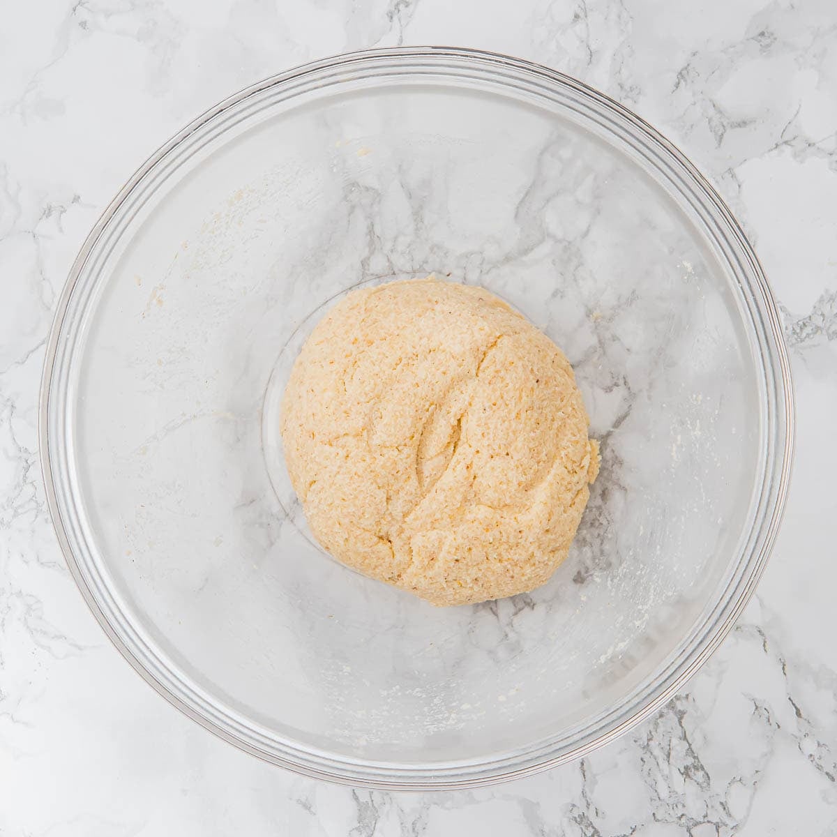 A round ball of tortilla dough sits in a clear glass mixing bowl on a white marble countertop.