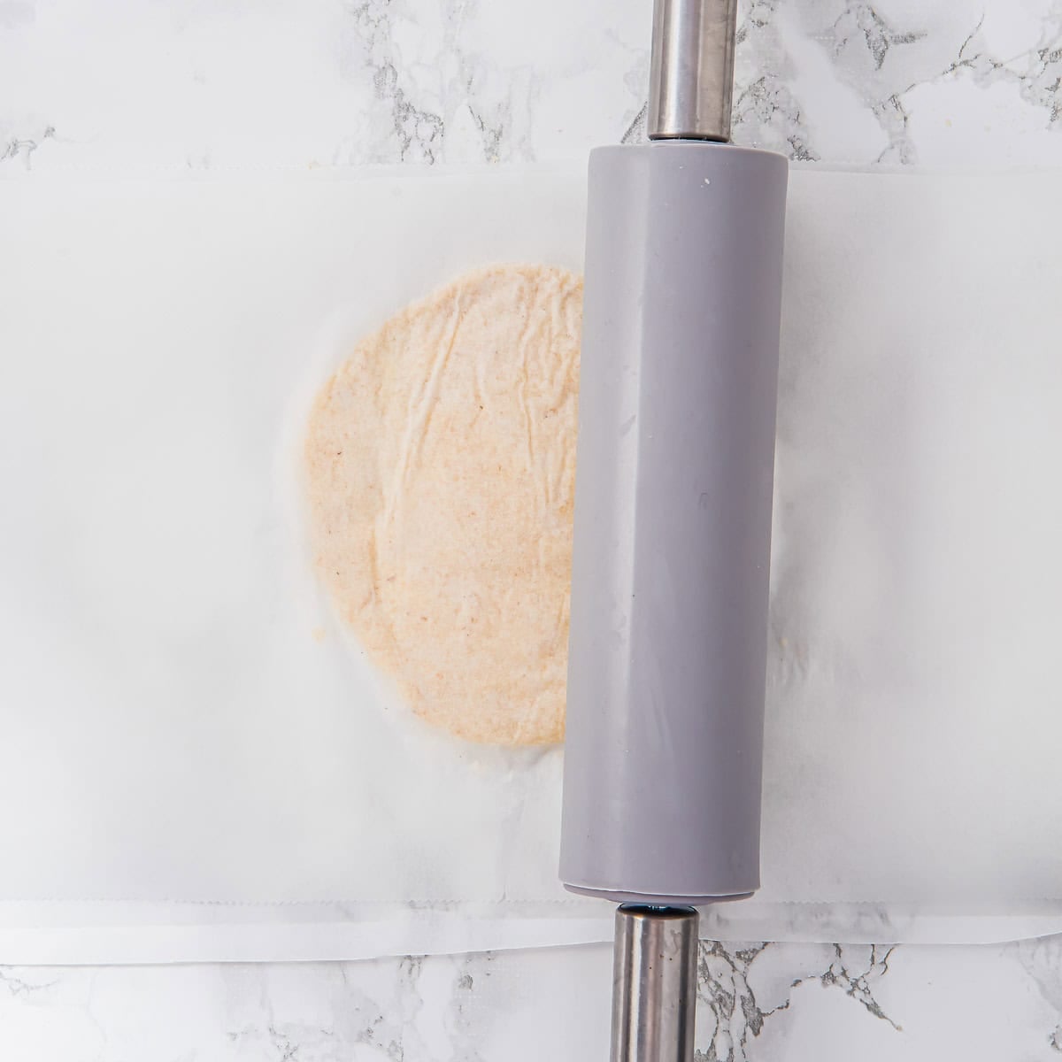 A ball of almond flour tortilla dough is being flattened under a sheet of parchment paper with a gray rolling pin on a marble surface.