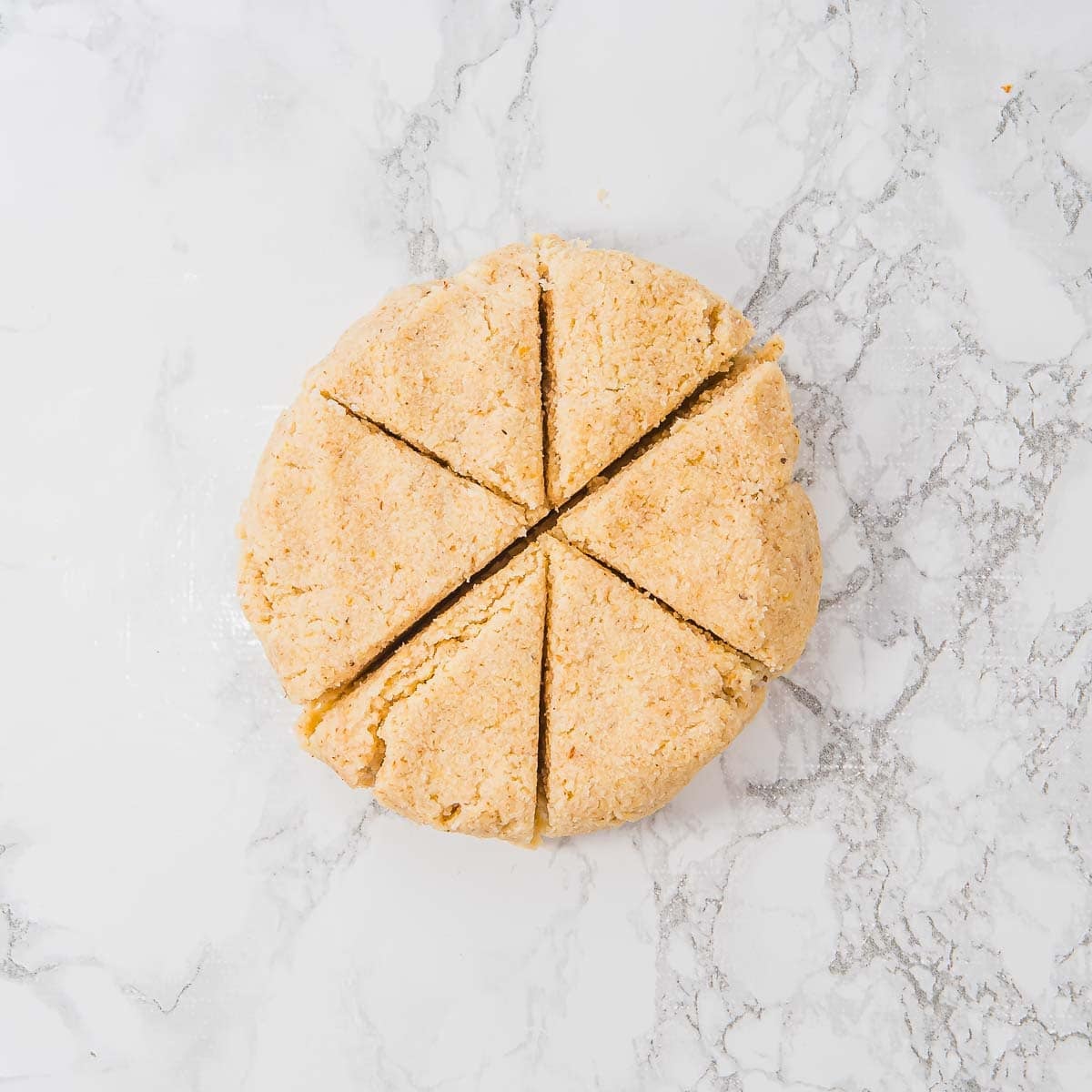 A round disk of tortilla dough cut into six wedges sits on a white marble surface, ready to be baked.