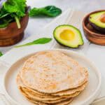 A stack of almond flour tortillas on a white plate, with a halved avocado, a bowl of mixed greens, and spinach leaves on a light-colored surface in the background.