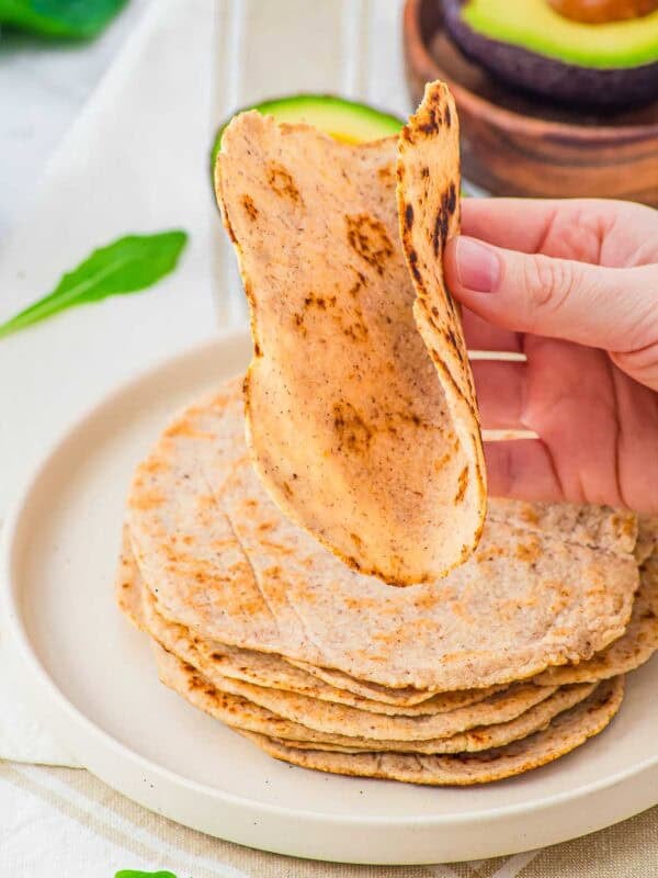 A hand lifts a homemade almond flour tortilla from a stack of tortillas on a white plate, with a sliced avocado and green leaves in the background.