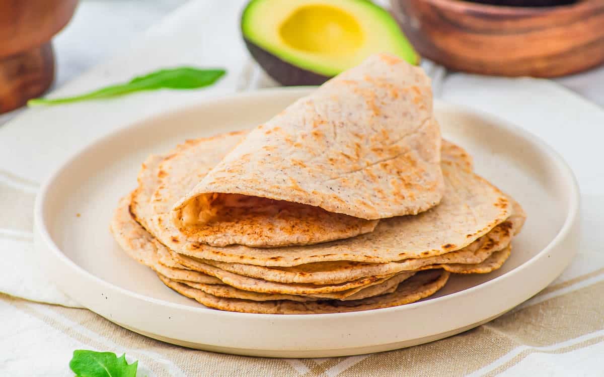 A stack of gluten-free almond flour tortillas is served on a white plate, with one tortilla folded on top. An avocado and leafy greens are visible in the background.