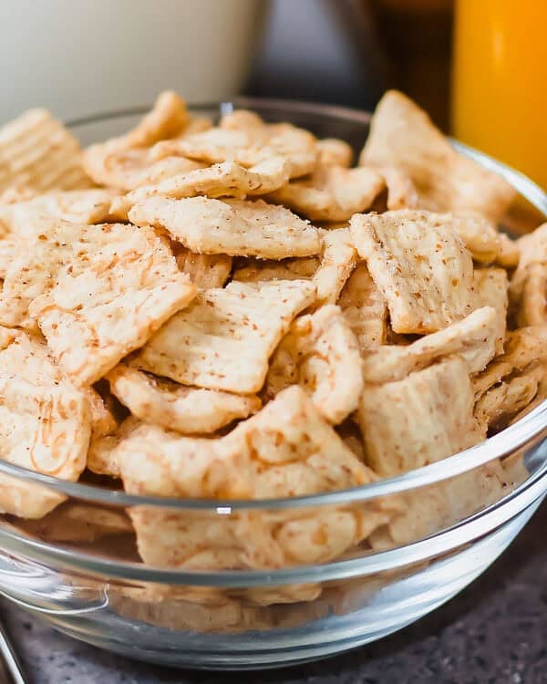 A clear glass bowl filled with square-shaped breakfast cereal pieces sits on a gray surface, with a spoon, a glass of orange juice, an apple, and a cup of milk blurred in the background.