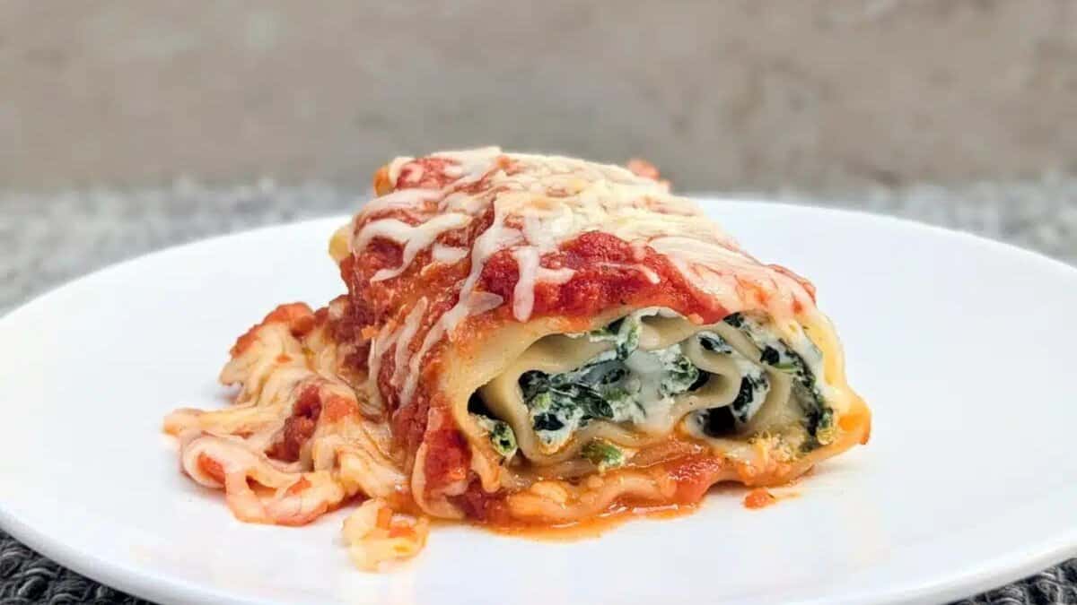 A single serving of spinach and ricotta lasagna roll topped with melted cheese and tomato sauce sits on a white plate against a neutral background.