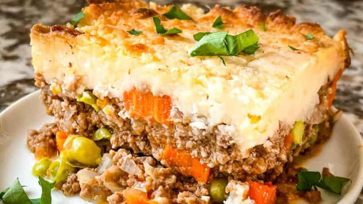 A slice of shepherd’s pie featuring layers of seasoned ground meat, peas, carrots, and mashed potatoes, garnished with chopped parsley on top.
