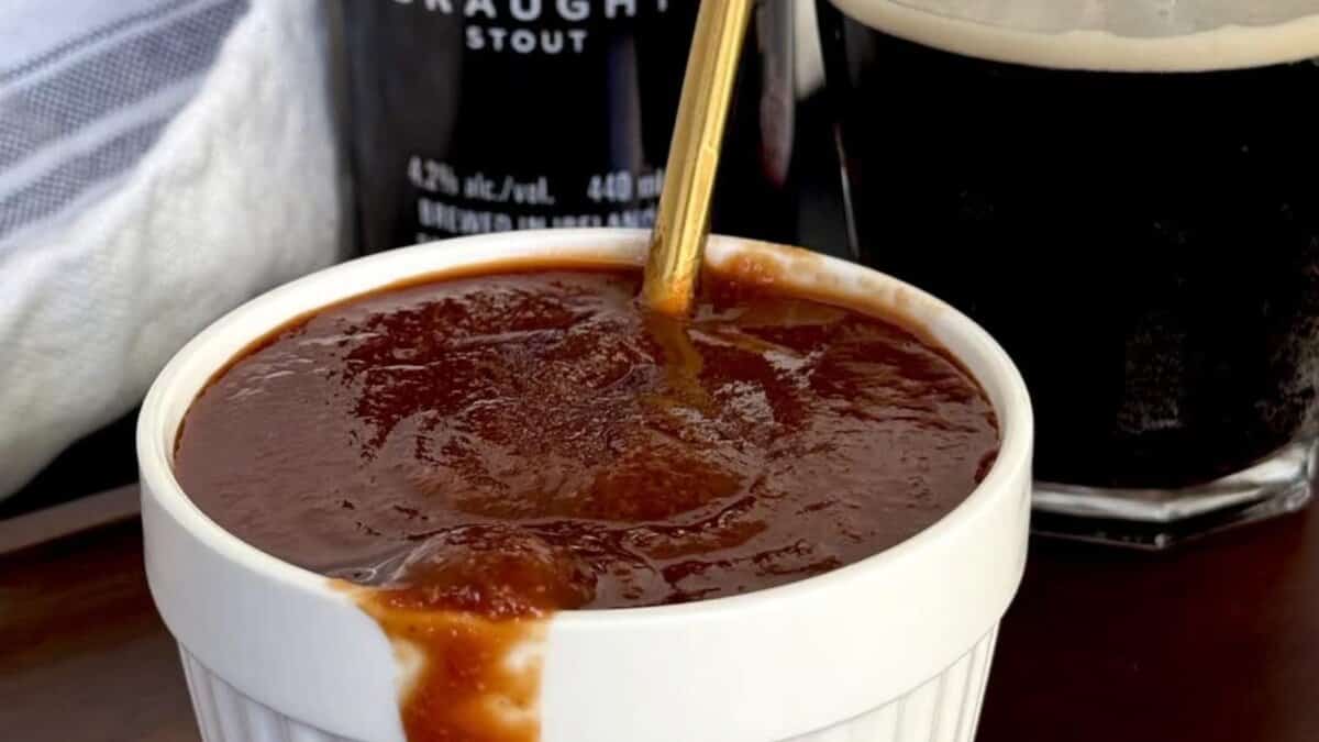 A white ramekin filled with thick, brown barbecue sauce sits on a table, with some sauce spilling over the edge and a gold spoon inside. A glass of dark stout beer is in the background.